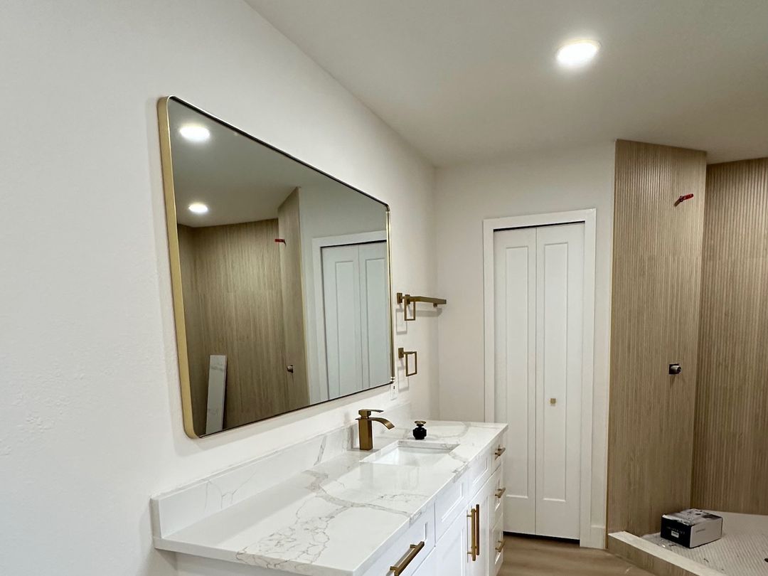 A bathroom with a white vanity, a stone top and a gold mirror.