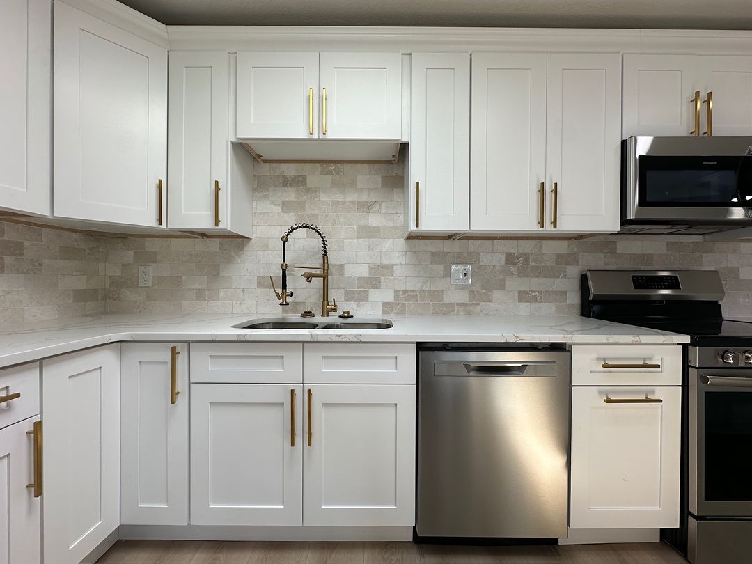 A white shaker kitchen with gold pulls, a stone backsplash and stainless appliances.
