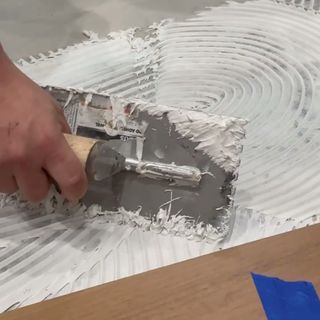 A notched trowel spreading adhesive on a concrete slab next to installed hardwood.