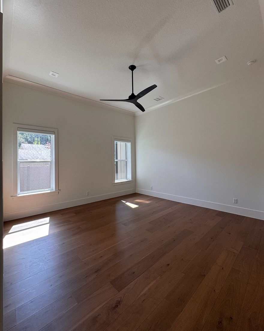 A bedroom with two windows, a black ceiling fan and sun on a hardwood floor.