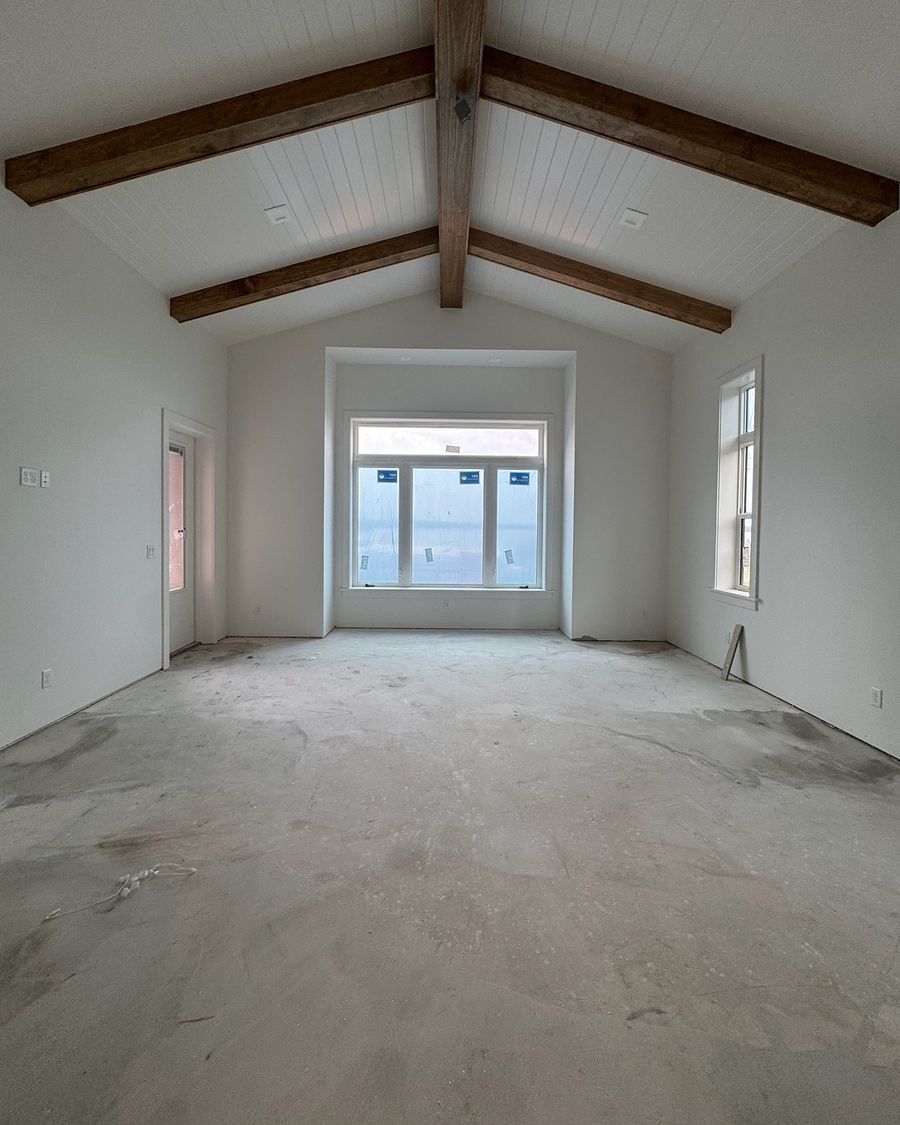 A bedroom with wood ceiling beams on day one: a bare concrete slab under a tall window.