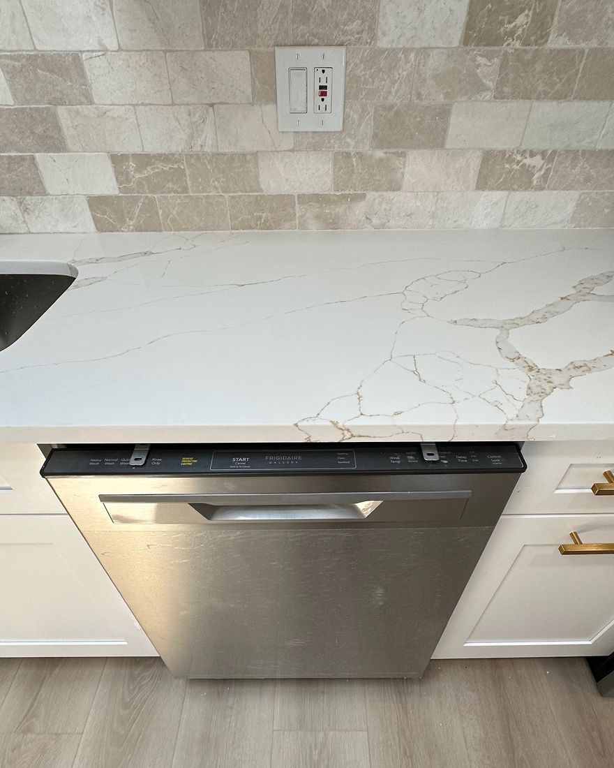 A white stone countertop with a stone tile backsplash over a stainless dishwasher.