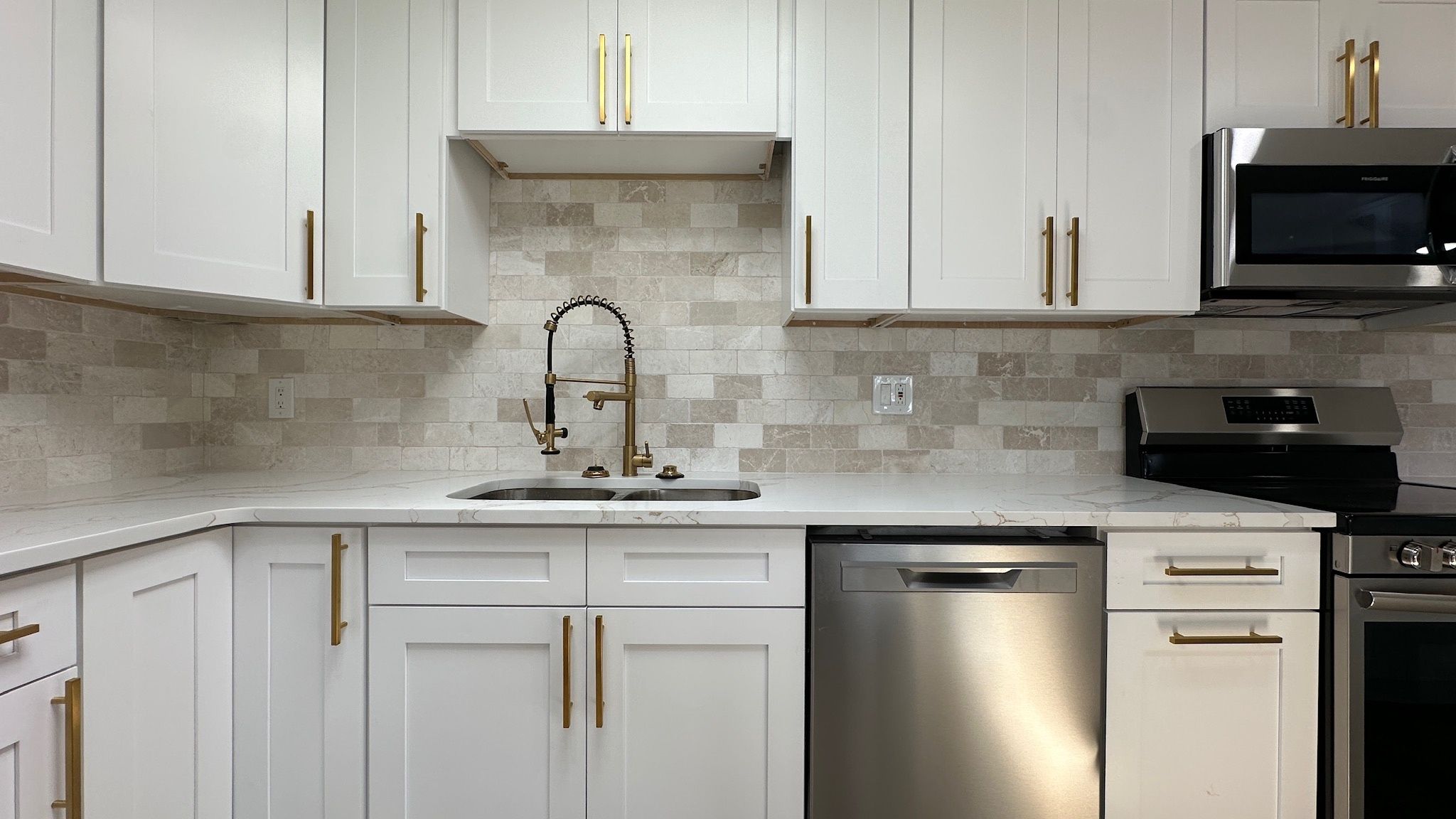A white shaker kitchen with gold pulls, a stone backsplash and stainless appliances.