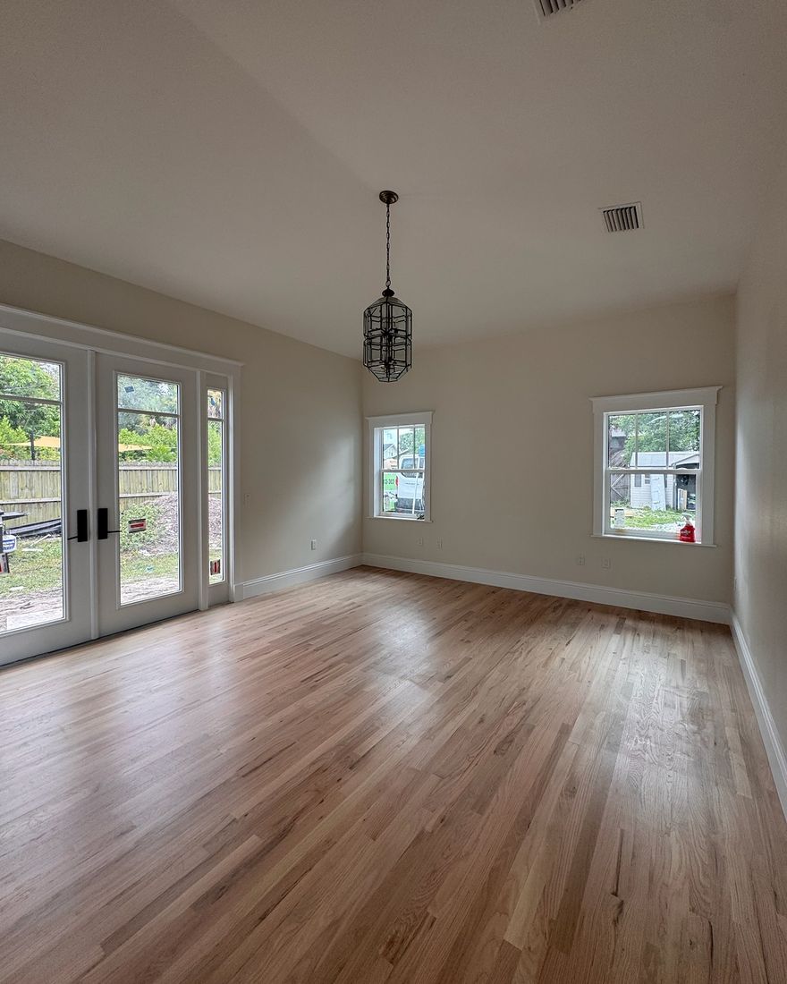 An empty living room with a natural hardwood floor, a pendant light and two windows.